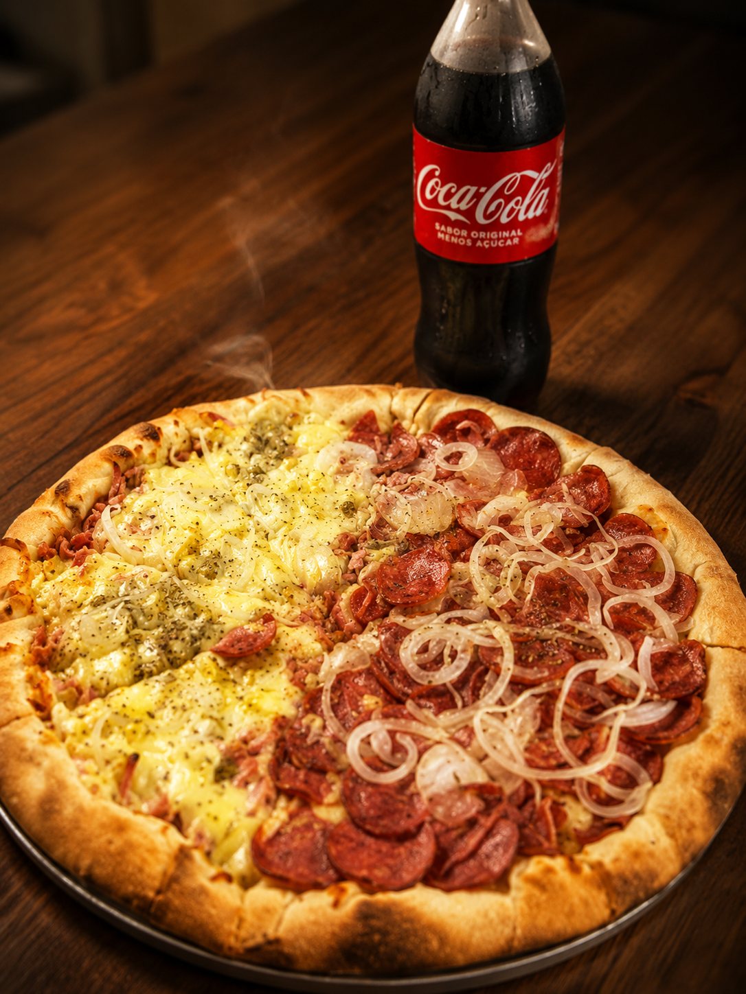 Duas pizzas meio a meio servidas na mesa de madeira da Bonna Pizza, com uma Coca-Cola ao fundo